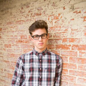 Trendy young man in glasses, plaid shirt, posing confidently against a brick wall.