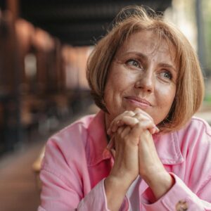Senior woman with short brunette hair and pink shirt, hands clasped, looking thoughtful outdoors.