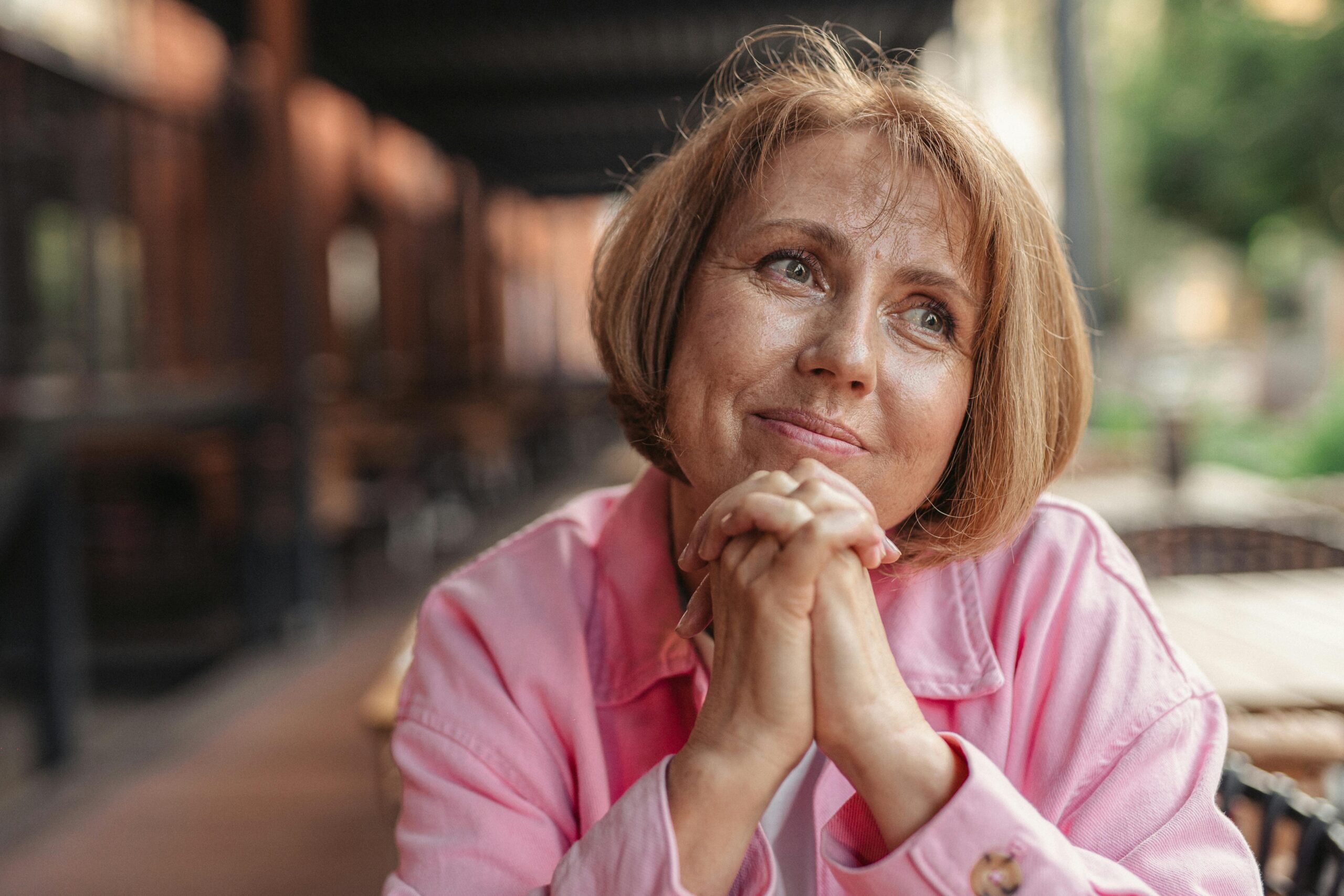 pexels photo 8972312 8972312 Senior woman with short brunette hair and pink shirt, hands clasped, looking thoughtful outdoors.